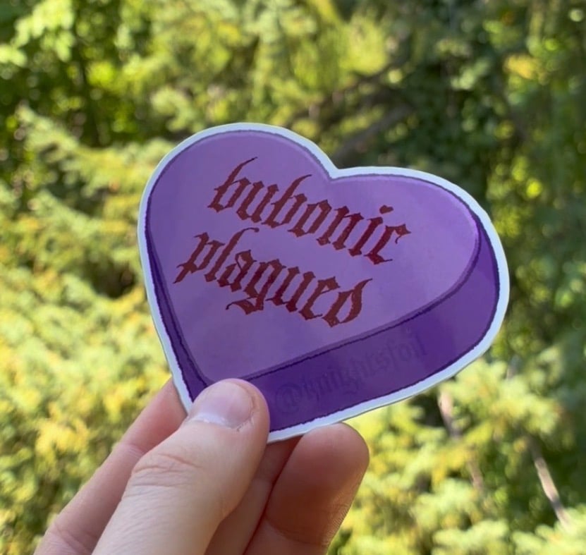 Photograph of my hand holding a sticker shaped like a purple heart. The handlettered text on the heart reads "bubonic plagued" in blackletter. The background appears to be blurry greenery.