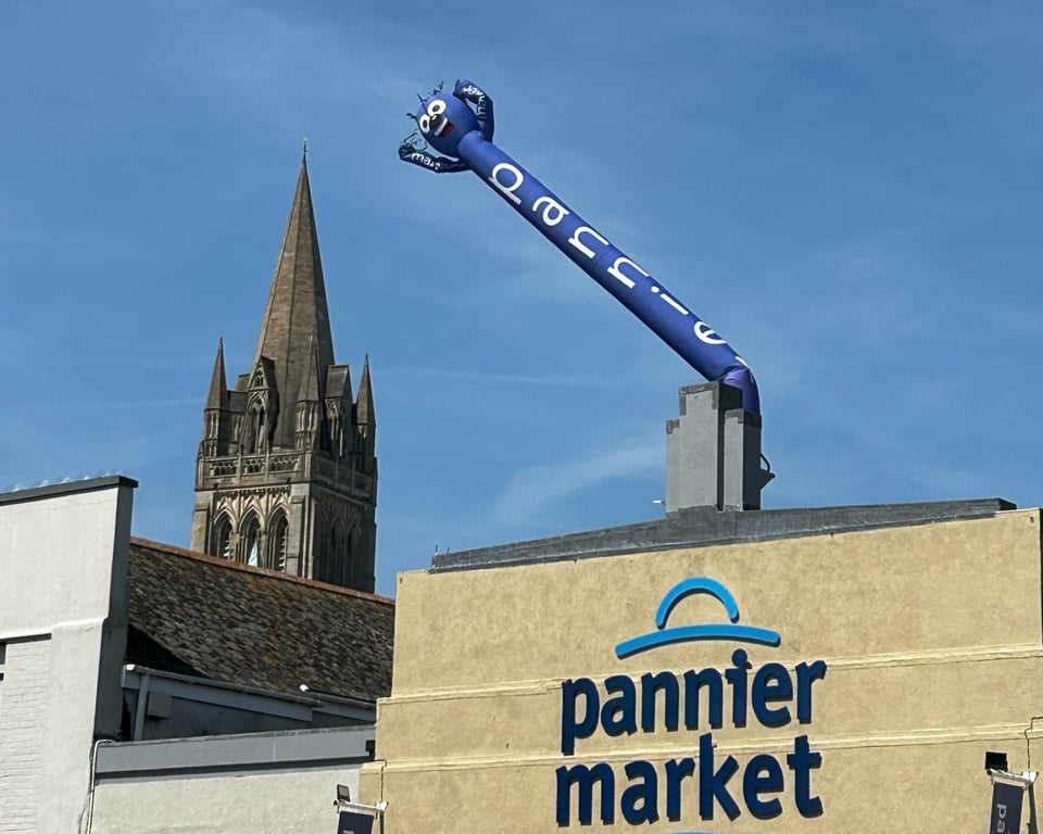 The tall spire of the Cathedral in the background is loomed over by a blue inflatable stylized man with 'pannier' printed on his body and 'market' on his arms. He is grinning madly, and emerging from behind a simple stepped facade with an Art Deco detail. It's not exactly stylish.