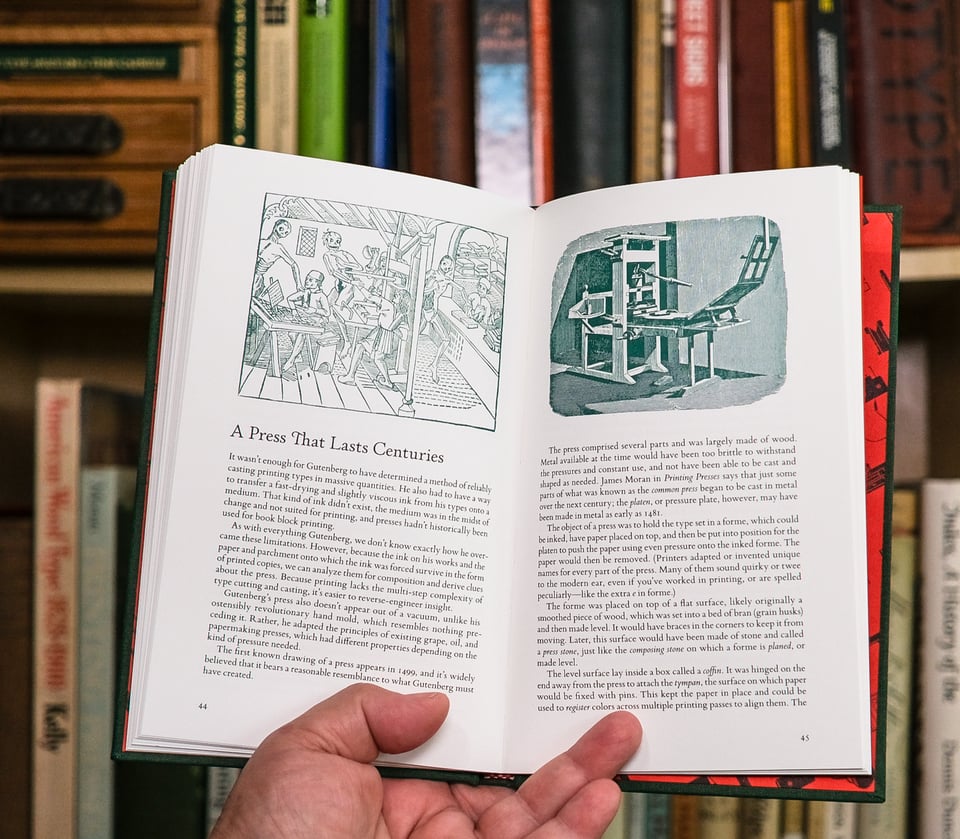 Spread of book with green illustrations at top and text below with hand holding it open in front of a slightly out of focus bookshelf of type and printing books.