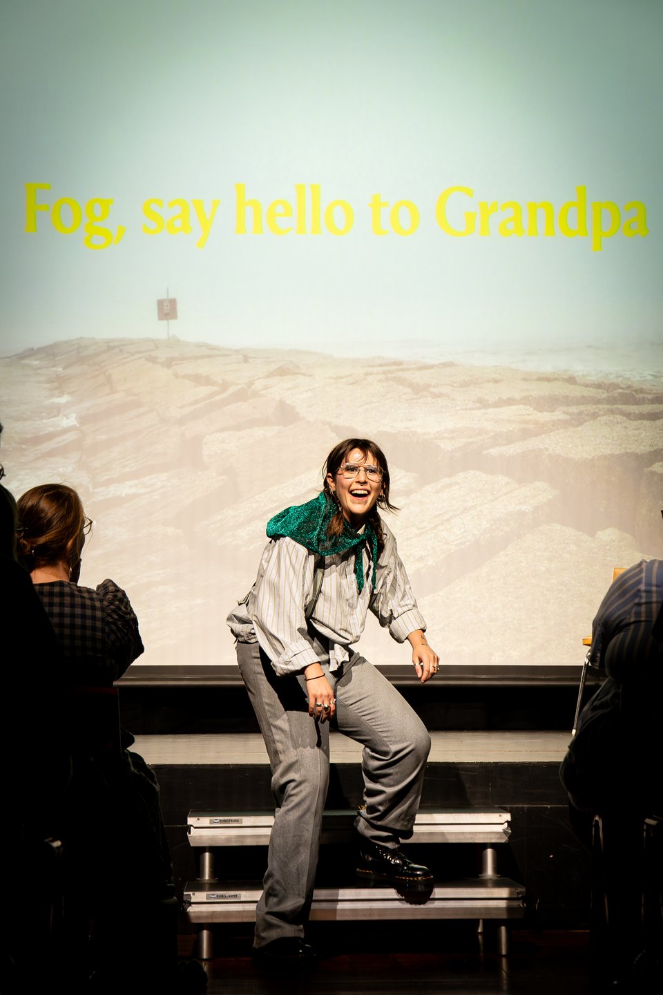 Mog as old man steps forward into the crowd with a befuddling smile. Behind them the screen shows a rocky jetty and bright yellow text saying 'Fog, say hello to Grandpa'