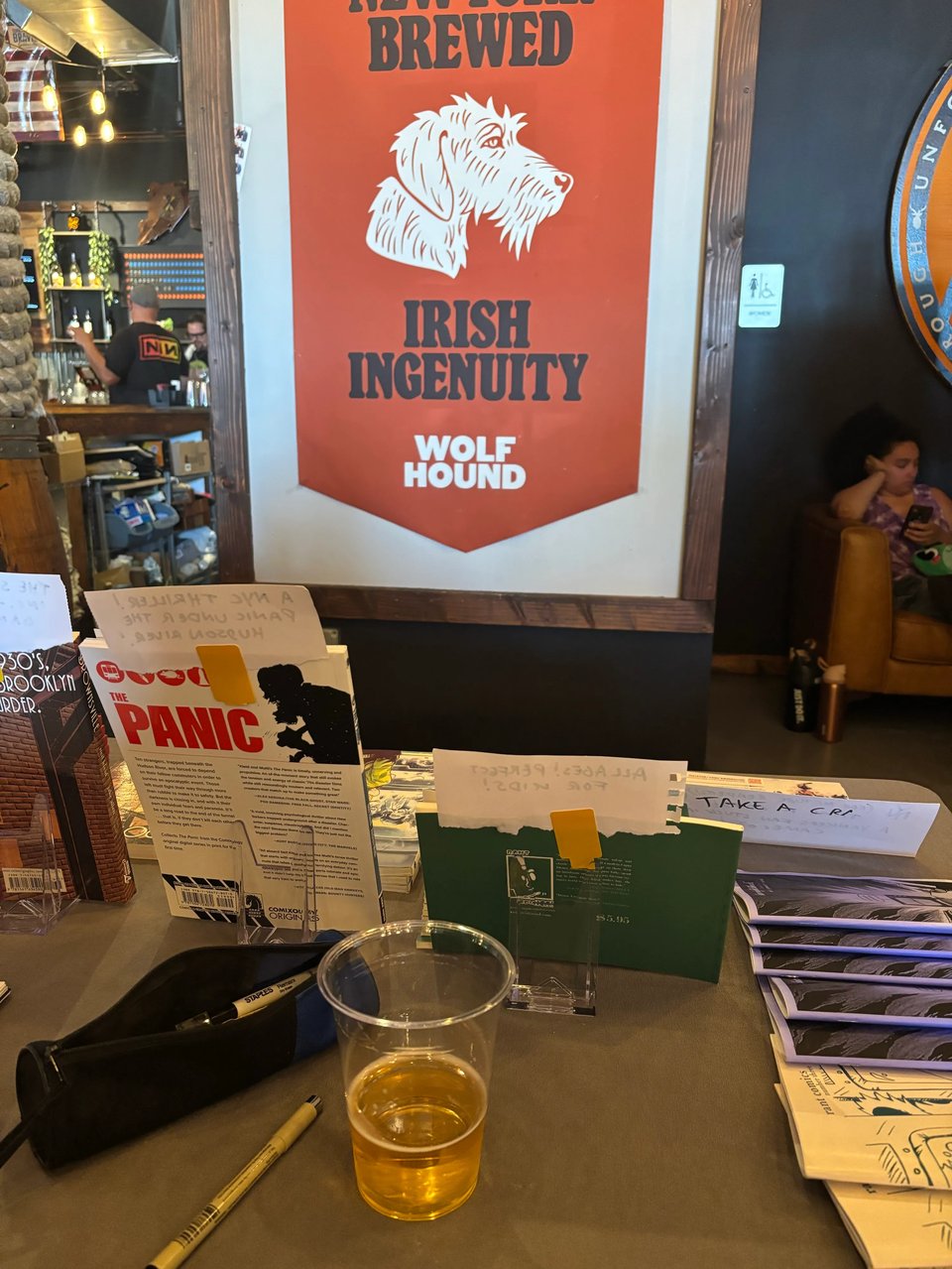 a picture from behind neil's table setup at staten island comics fest, with a half empty beer and some of his books for sale and a wolf hound beer poster showing a wolf hound dog (?) that says New York brewed, Irish ingenuity
