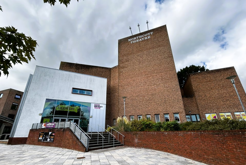 A theatre made up of three large masses. The central block includes the fly tower and is plain brick, with a lower block also of brick to its right. To the left is a white rendered block with double height doors in it, which is the entrance.