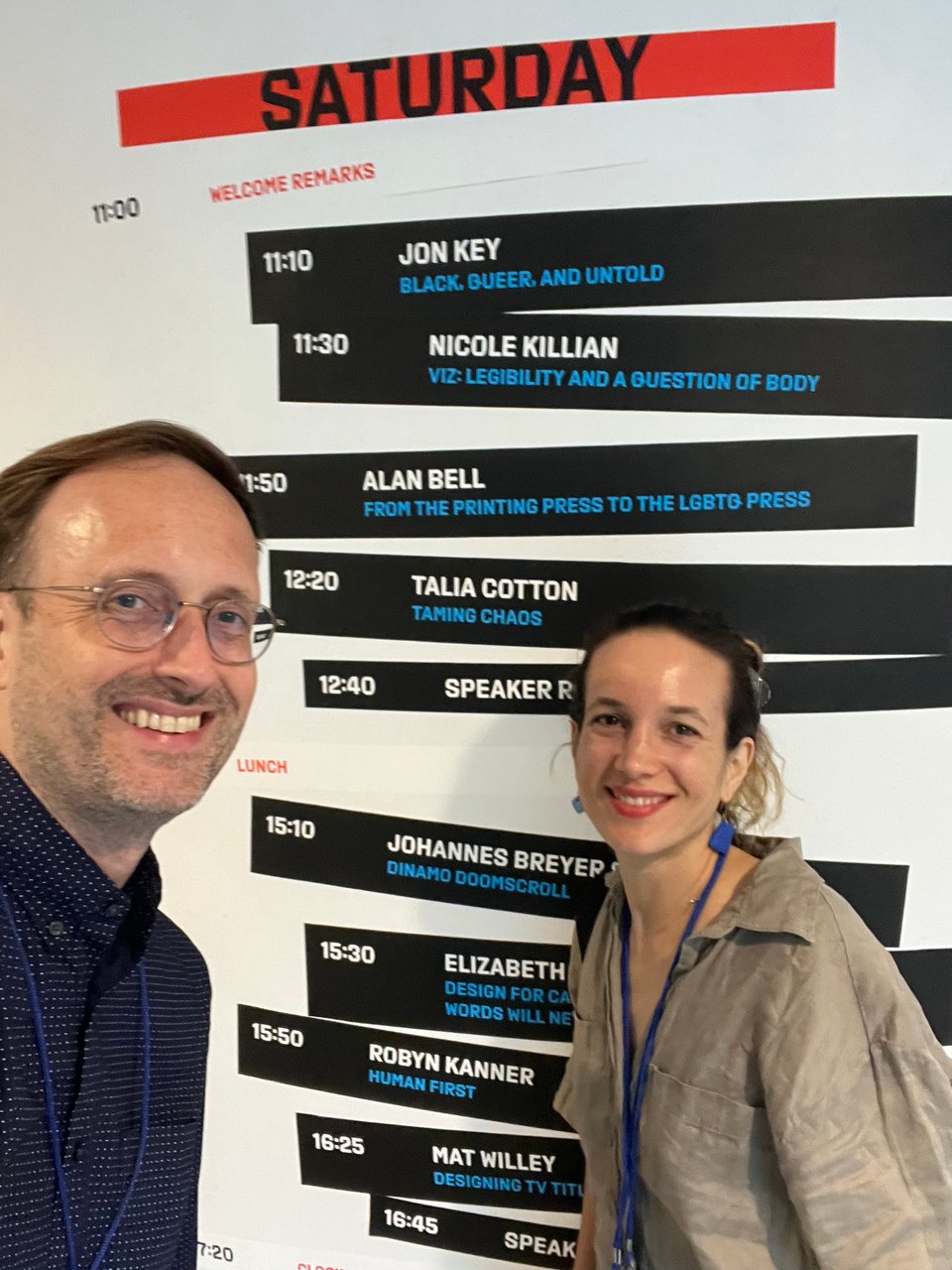 Man with glasses and woman with a blue lanyard smile awkwardly in front of the conference schedule, set in Export