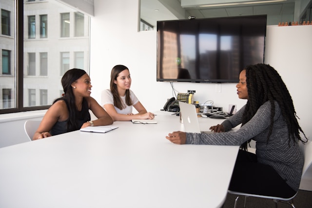 Foto de três mulheres num ambiente profissional sentadas ao redor de uma mesa, duas de um lado e uma de outro, debatendo alguma coisa.