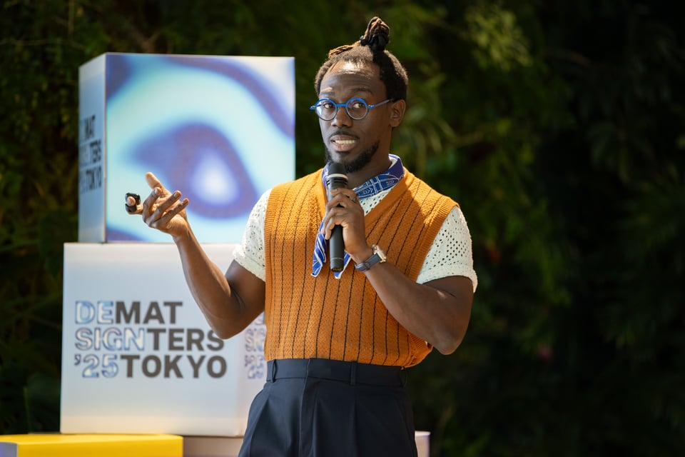 A man in blue-rimmed glasses and a sweater vest speaking on stage.