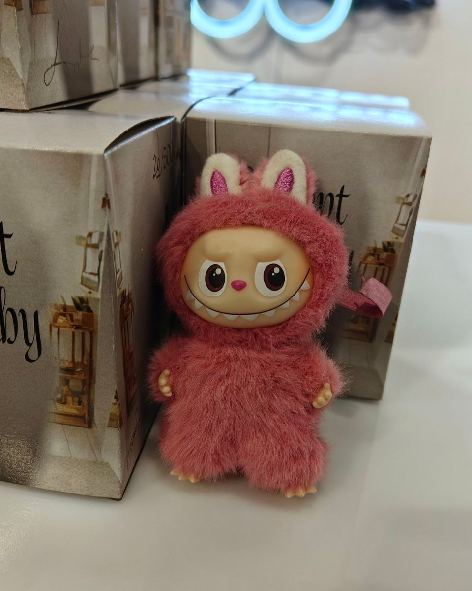 A photo of mini pink Labubu in front of some small product boxes, which are grey toned and say "Prompt Baby" on them. They are the exact same size as the popmart boxes