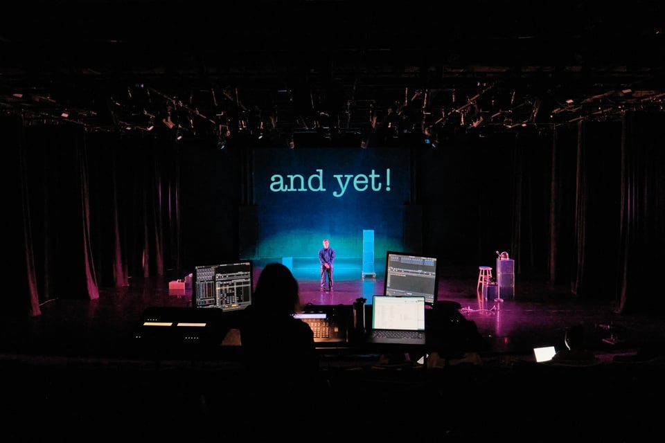 a shot from deep in the seats of the theater at the Dance Center at Columbia. a person's head is in silhouette at the lighting book. Nora stands on the stage. the words