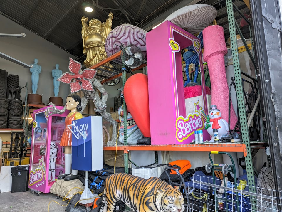 Shelves include a giant Barbie Doll, a tiger statue, a doll, a big thing saying let's GLoW SF, a stormtrooper from Star Wars, and other junk