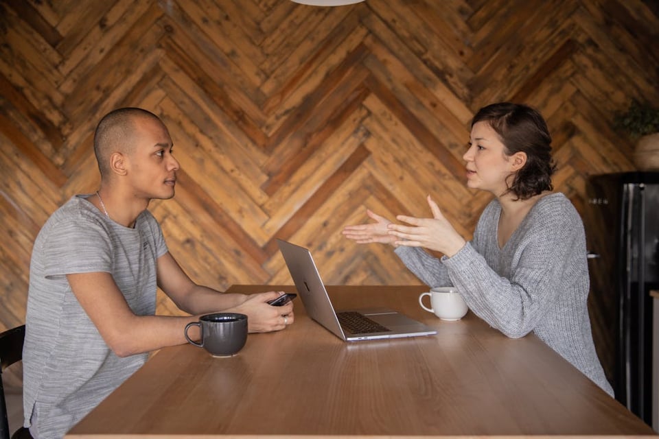 Two people sitting facing each other at a wooden table, talking with close concentration to each other. A young woman with short dark hair is wearing a gray sweater and gesturing with her hands towards a man who is holding a phone and looking at her intently, wearing a gray T-shirt. The woman has an open laptop in front of her and a white mug of coffee; the man has a black mug of coffee in front of him.