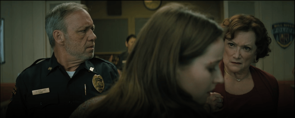 Police Chief Collins, an older white man with mostly grey hair and a five o'clock shadow in his uniform, and Mrs. Collins, an older white woman with red-brown short curls in a red outfit, flank either side of Brynn. Brynn's head is turned to the side after Mrs. Collins has spit in her face. Behind them, the two officers at the front desk watch silently.