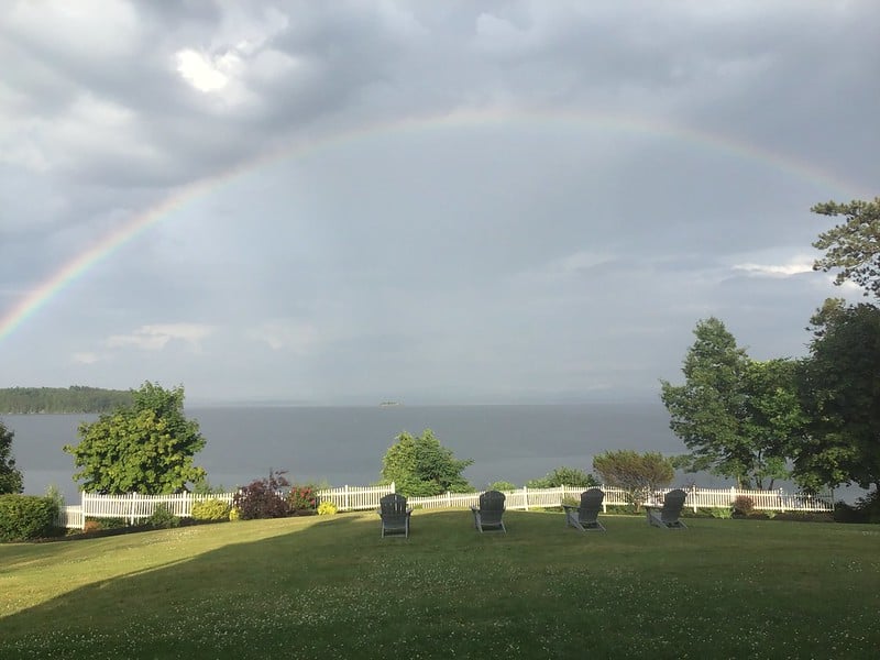 Rainbow over a river with Adirondack chairs in the foreground, and a grassy field