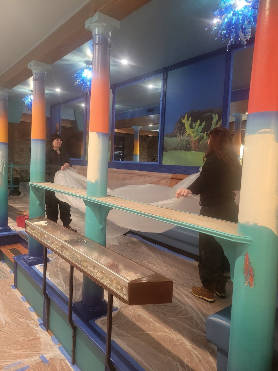 Image of the gallery seating, with coral reef paintings on the wall panels and sunset ombre colors on the pillars. Two friends lay down painting tarps.