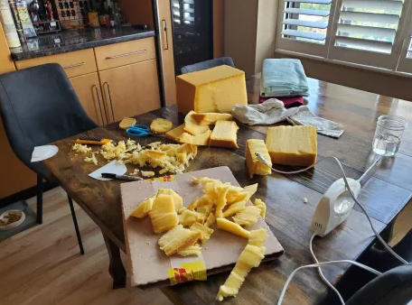 An extraordinarily messy dining room table. Large blocks and small shavings of parmesan-esque foam litter a table. An electric bread knife can be seen nearby. There are scissors, a utility blade, a sharpie, and — inexplicably — two semicircles of plastic cut from a margarine container. It’s chaos, basically.