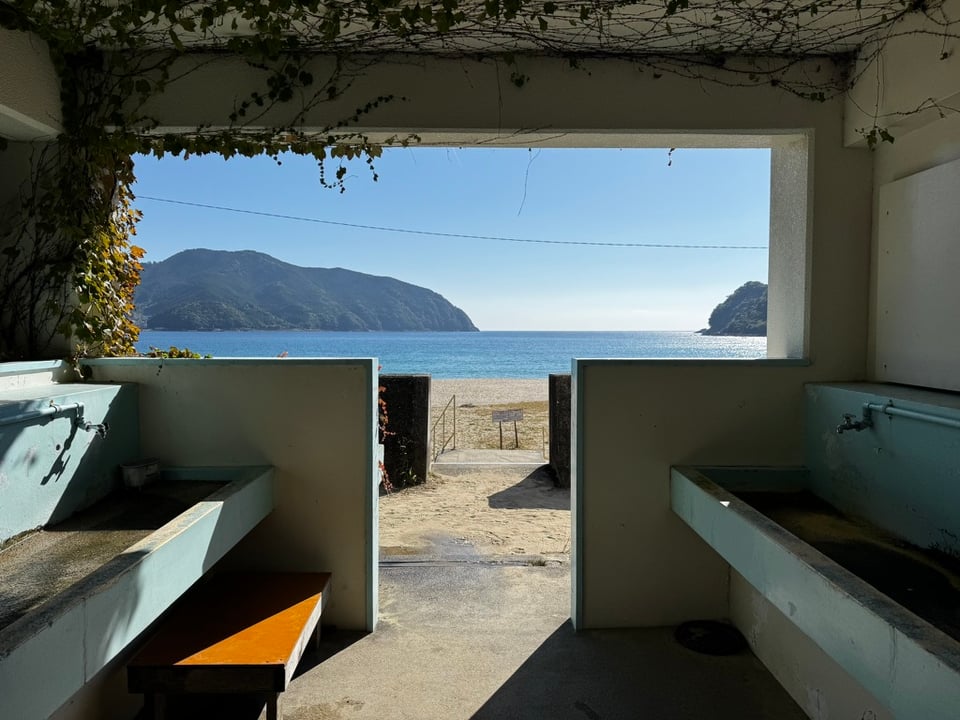 A view through a concrete facility with basic fixtures—sinks visible on either side—looking out towards a pristine coastal landscape. The framed vista shows a crescent sandy beach, calm turquoise water, and mountainous islands under a clear blue sky. Climbing ivy and vines frame the top of the opening. Power lines cross the view, and a small railing is visible on the beach access path. The stark architectural foreground contrasts dramatically with the serene natural seascape beyond.