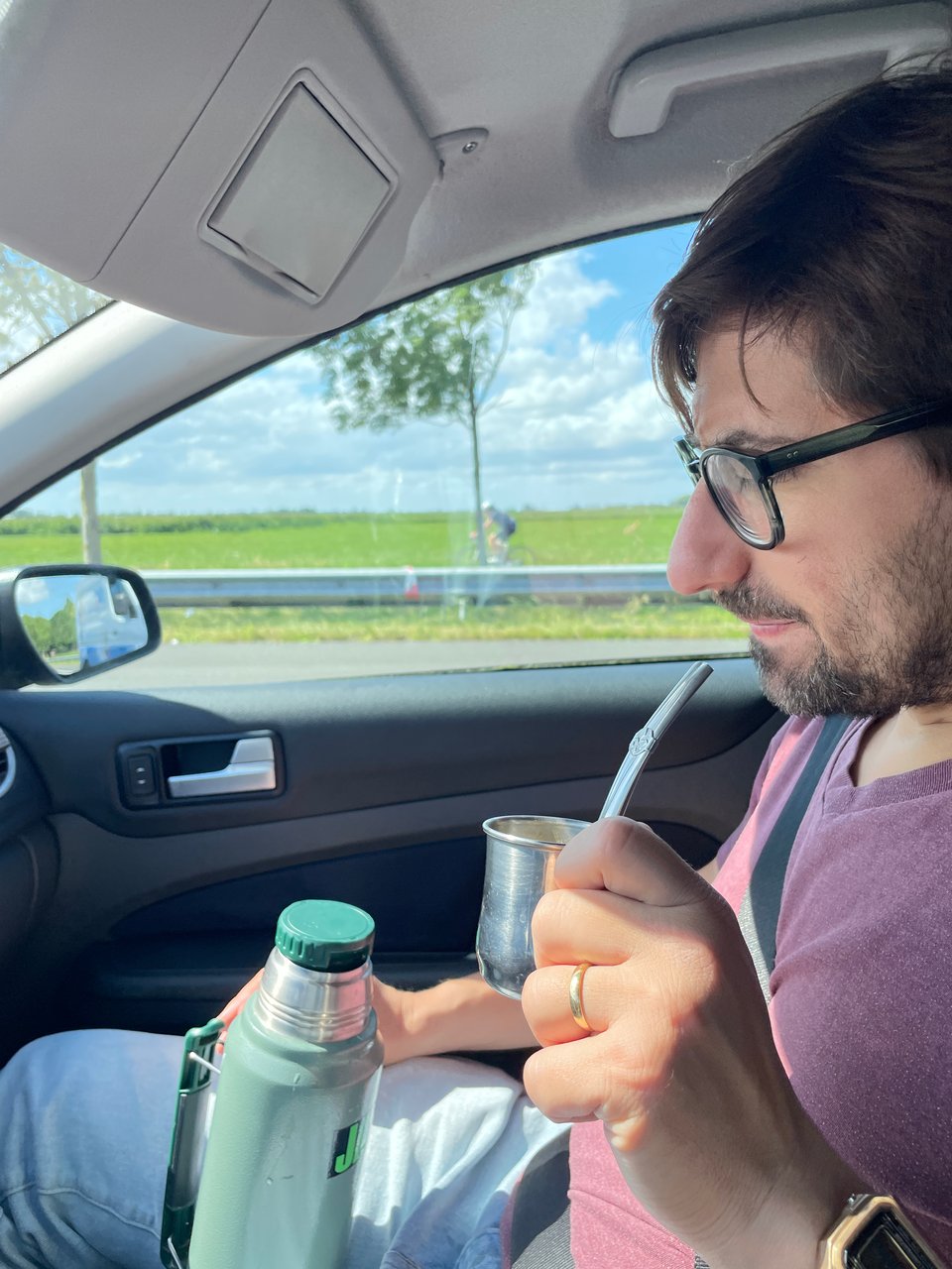 A man in a purple t-shirt sits in a passenger seat and drinks yerba mate from a small tin cup with a bombilla and has a large green thermos between his legs.