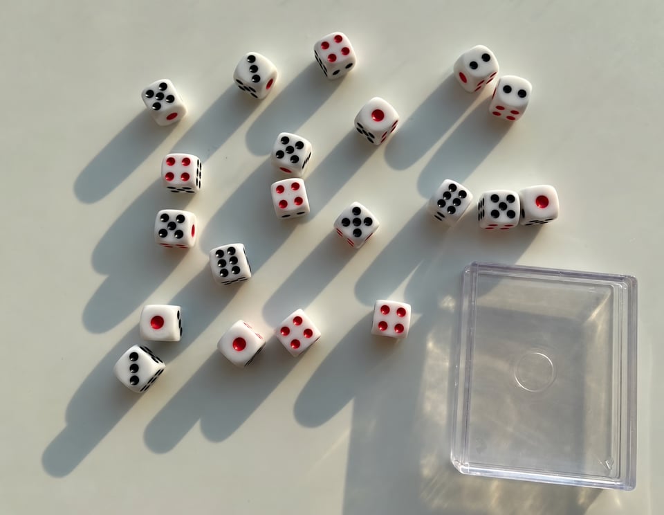A set of 20 small dice in the evening light. They are white with red and black etchings. A small clear plastic box is next to them.