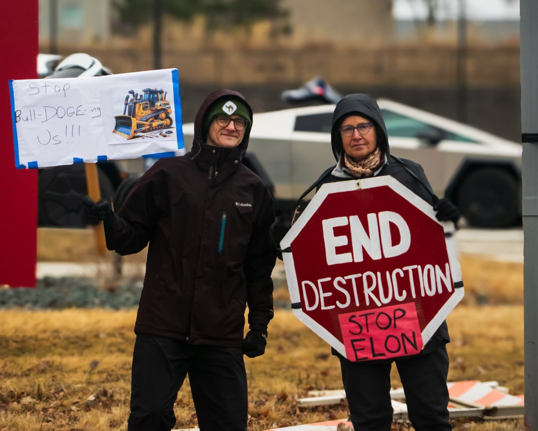 a couple stands in front of a Tesla dealership. There is a cybertruck in the background being shown to a potential buyer. The man on the left is smiling proudly and holding up a sign that says Stop BullDOGEing us!!! It has a picture of a bulldozer on it. The woman, right, looks determinedly into the wind with a large stop sign that says End Destruction, Stop Elon.