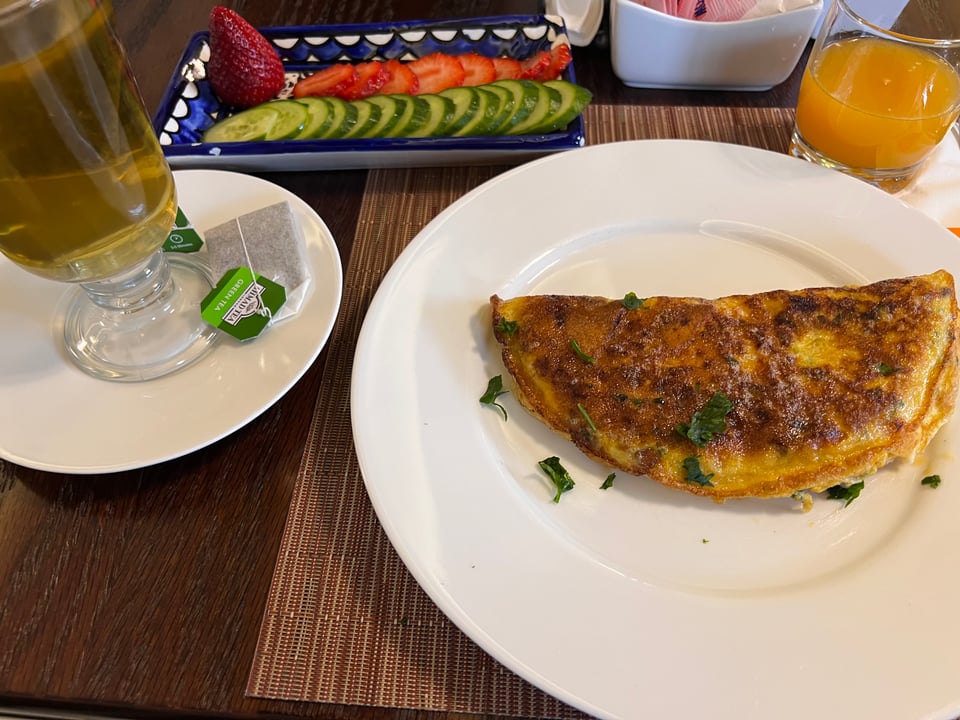 A white plate holding a folded omelet, a cup of tea, fresh orange juice in a glass, and a blue and white plate with sliced cucumbers and strawberries
