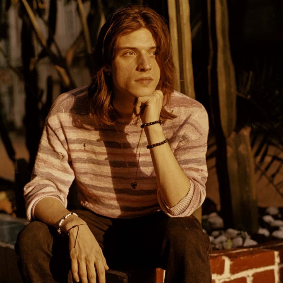 A young man with shoulder-length hair and a striped longsleeve shirt sits with his left elbow on left knee, resting his chin on his hand.