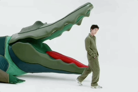Gif of Yibo in an olive-green fleecey jumpsuit, going on impossible fucking en pointe in a pair of sneakers in order to do an equally impossible slow graceful high kick — WHILE EN POINTE — in order to imitate the open red mouth of the giant green plastic Lacoste crocodile he is standing next to.