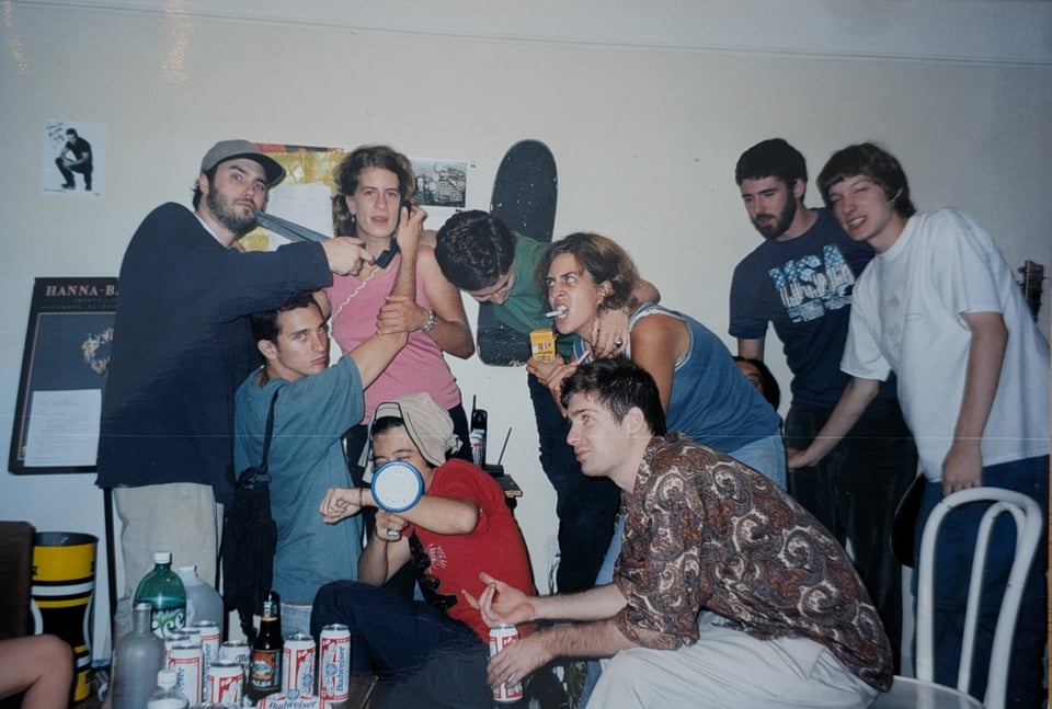 A group photo from some house part in 1998. Everyone poses in an unusual way. There is a phone, megaphone, duct tape, cigarettes being used as props. One man in the foreground holds a budweiser and isn't aware of the photo being taken. Two others lean in last minute. One woman has her head down like she's about to fall off the chair she is standing on.