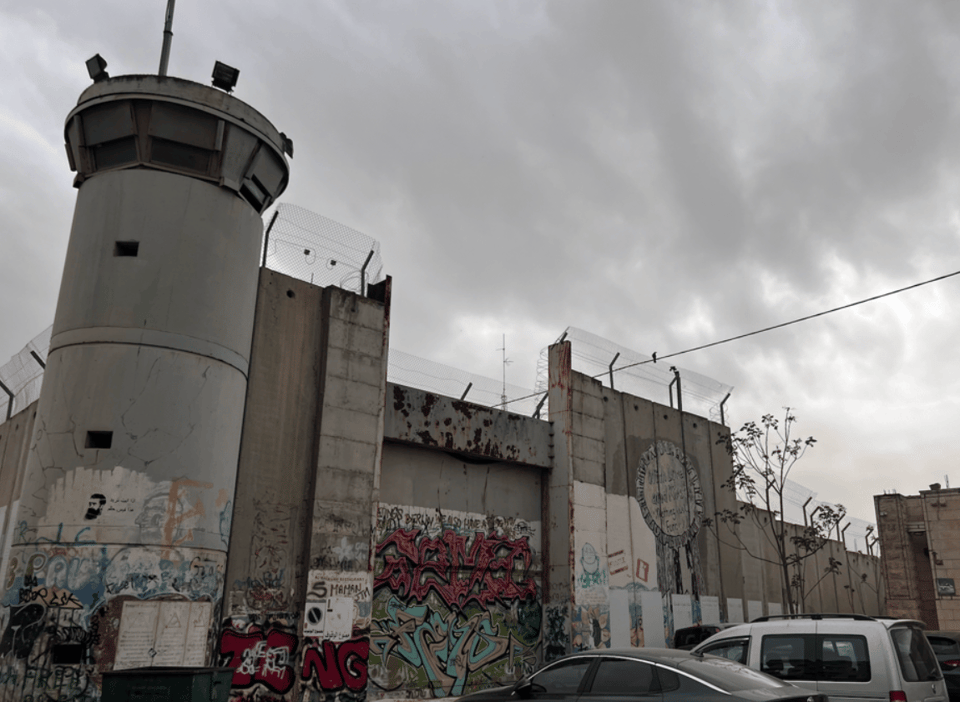 A concrete barrier with barbed wire at the top, and a military tower with 360 degree windows at the top. The wall is part of a parking lot, and has graffiti painted on it 