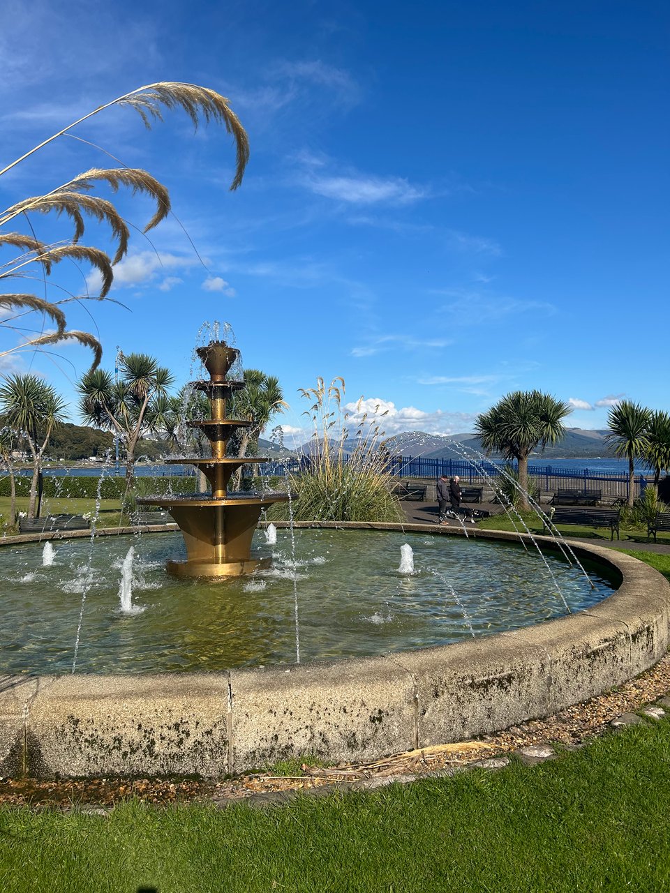 The Victorian fountain in the promenade gardens in Rothesay on the Isle of Bute. There are palm trees nearby and rounded hills in the distance. Image by Rowan Ambrose.