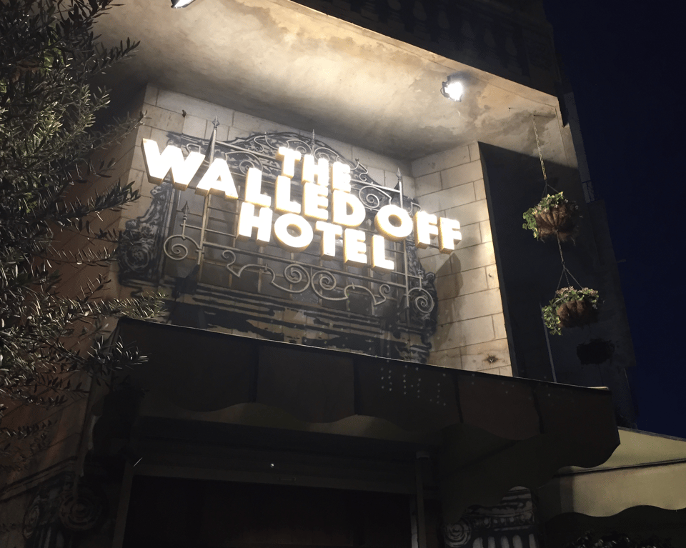 Nighttime photograph showing a hotel entrance with an illuminated  sign that says "THE WALLED OFF HOTEL". Decorative wrought-iron panels, stone columns, exterior lights, hanging plants, and dark awning frame doorway.