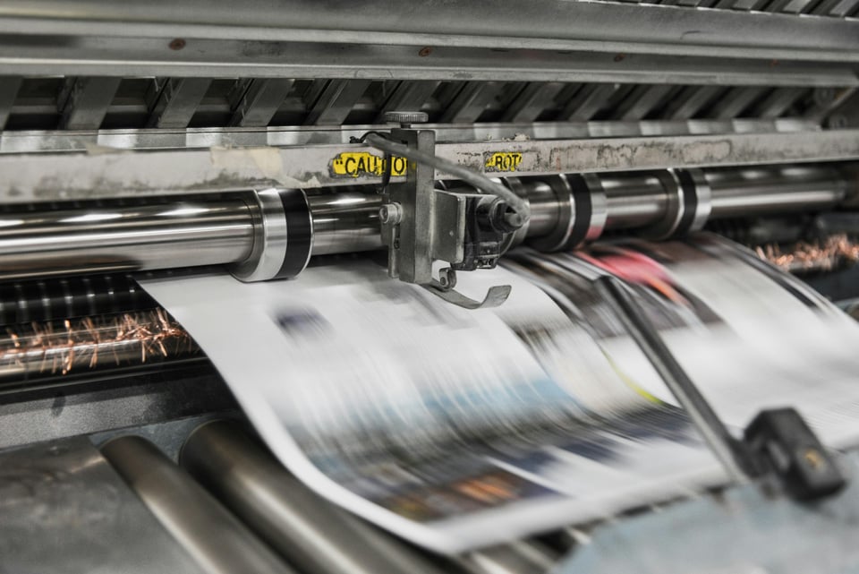 A newspaper printing machine in action. The paper, which is in motion, is a blur.  Photo by Bank Phrom on Unsplash.
