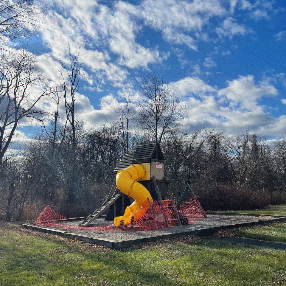 a bright yellow slide attached to a wooden playground remains fenced in by collapsing orange fencing - on the side of the slide you see a warning sign, apparently the playground is dangerous. below green green grass and above a beautiful early winter Catskill sky