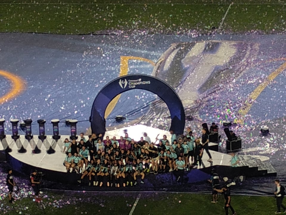 Gotham FC's team in black uniforms surrounded by coaches wearing sky blue jackets posing with the Concacaf W Champions Cup trophy on a stage a midfield.