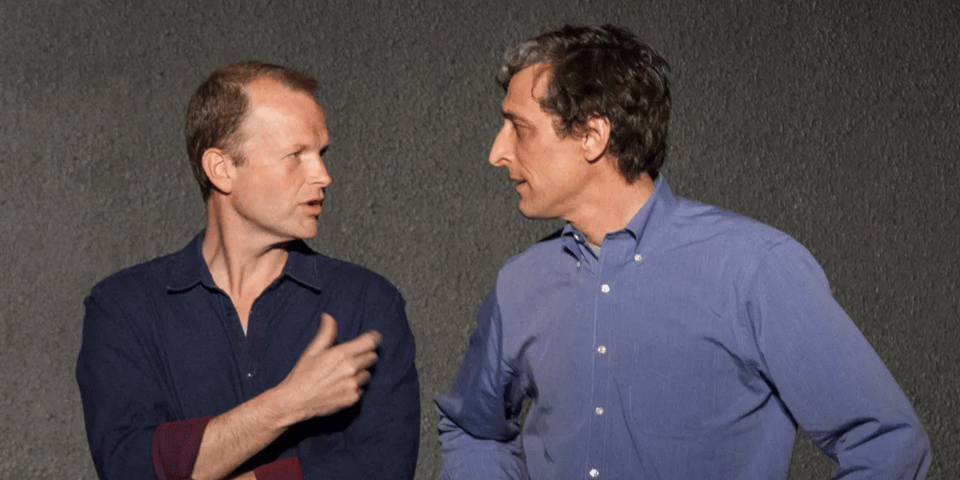 TJ & Dave, two white men in middle-age, stand looking at each other.