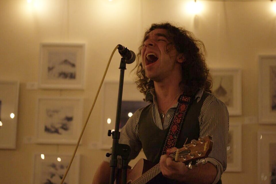 A person with wavy, shoulder-length brown hair wearing a buttondown shirt under a vest plays an acoustic guitar and sings into a microphone.