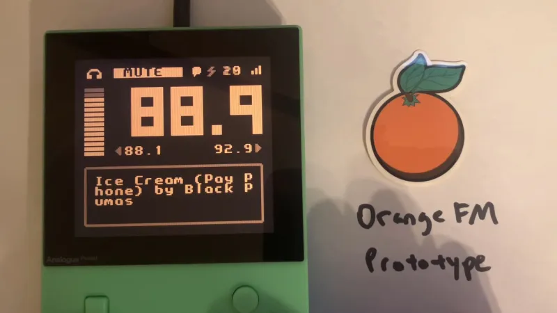 A photo of a green game boy with a jet black screen. On the screen is glowing large text, colored beige. The text shows the numbers 88.9, and beneath it you can read the title of the song currently playing: "ice cream (payphone) by black pumas". Next to the game boy is a die cut sticker in the shape of an orange, with a text written on the surface beneath it, "orange, FM prototype".