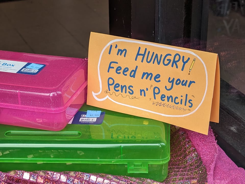 A display of plastic boxes to hold pencils, with a sign with a word balloon that reads: I'm HUNGRY! Feed me your Pens n' Pencils