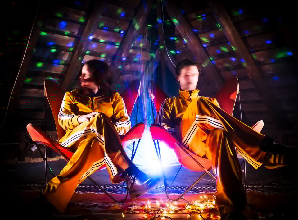 A woman and a man in matching yellow track suits sit side by side in red butterfly chairs. Each has their right leg crossed over their left knee, and their faces are blurred as if in motion.