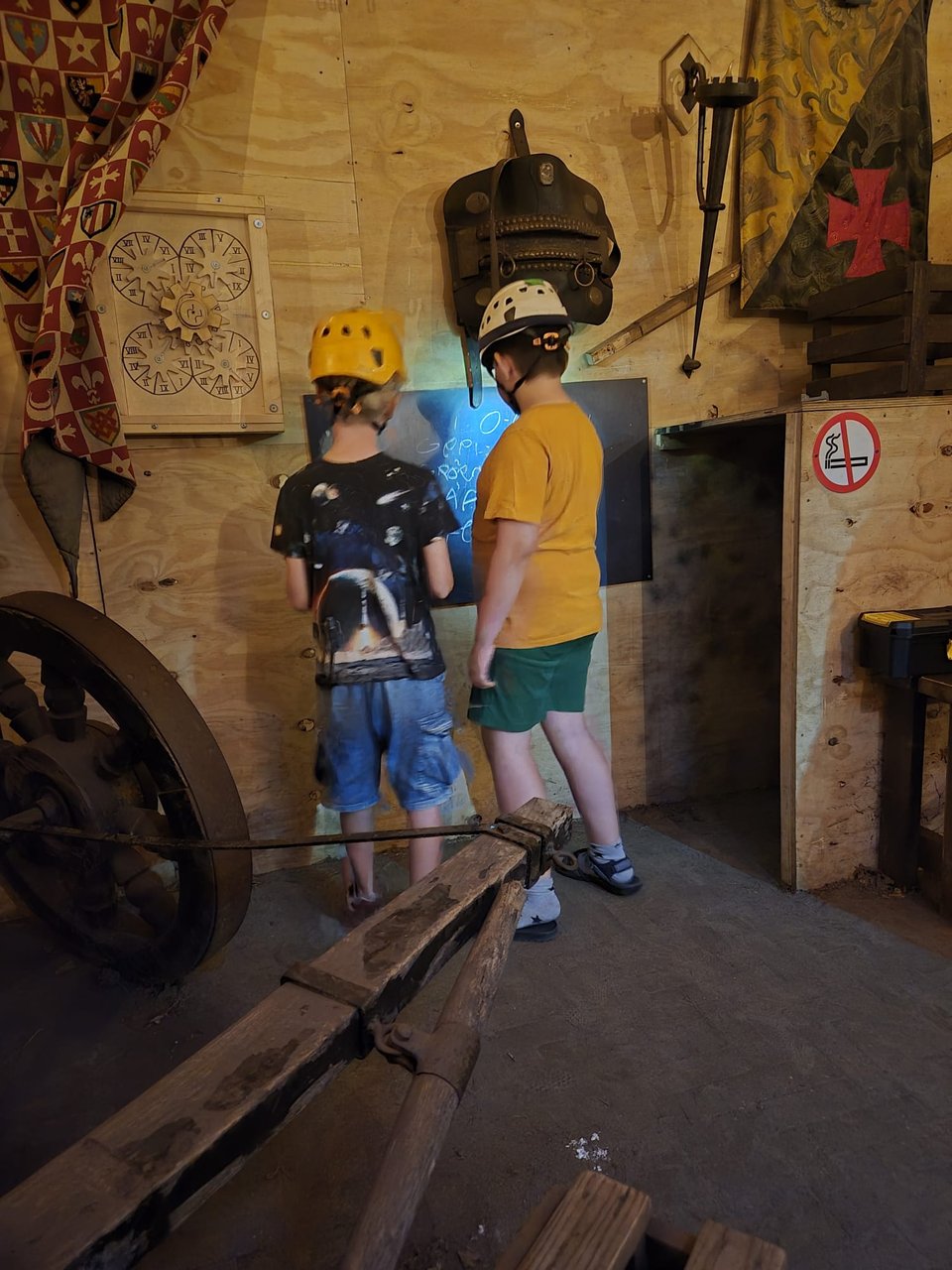 Two boys are trying to figure out clues in an escape room. They face a blackboard with notes scribbled on it. They are inside and wearing helmets with lights attached.