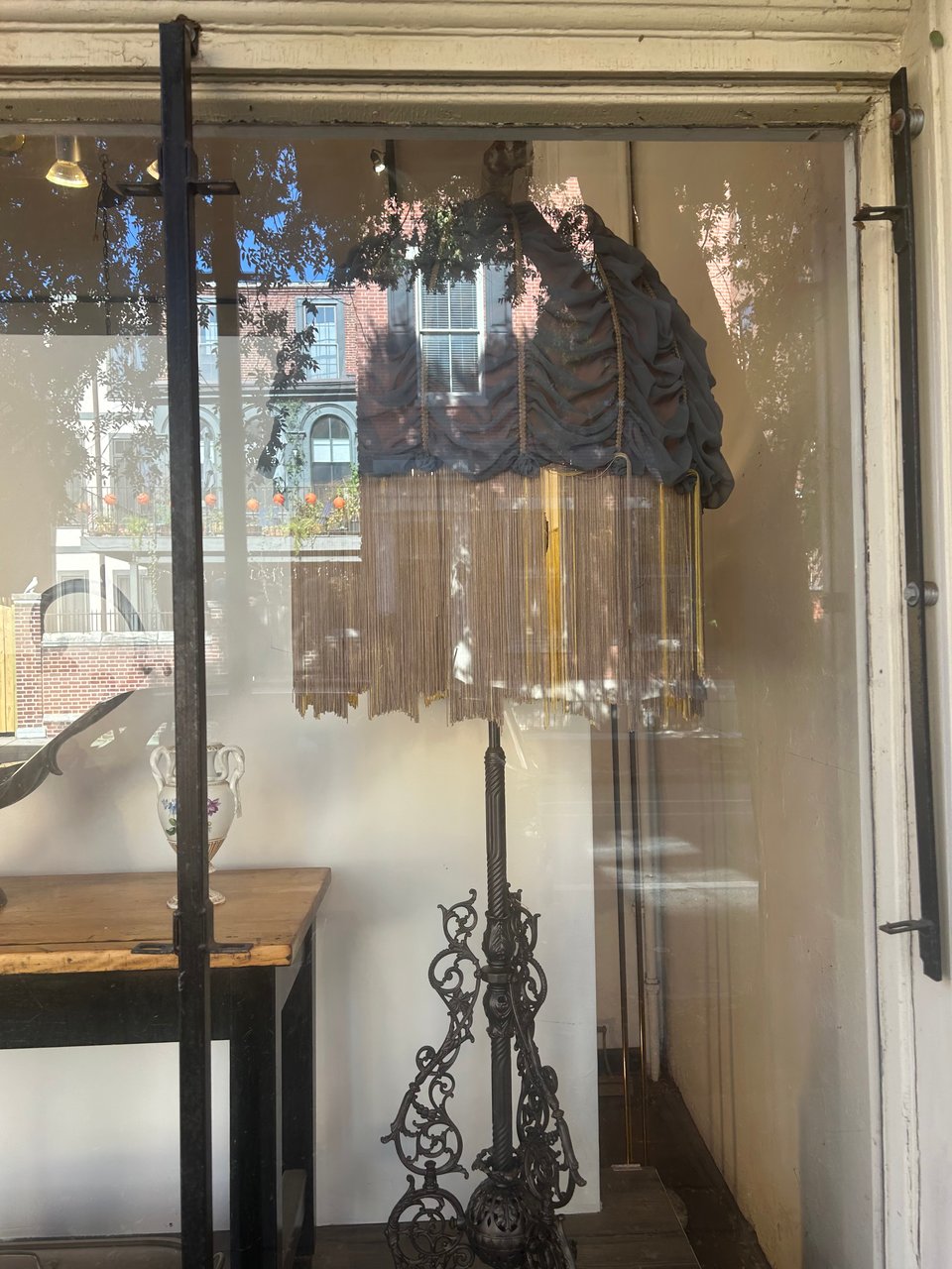 a photograph of a tall, baby blue fringed lamp with a gilded lamp base standing in the window of a shop.