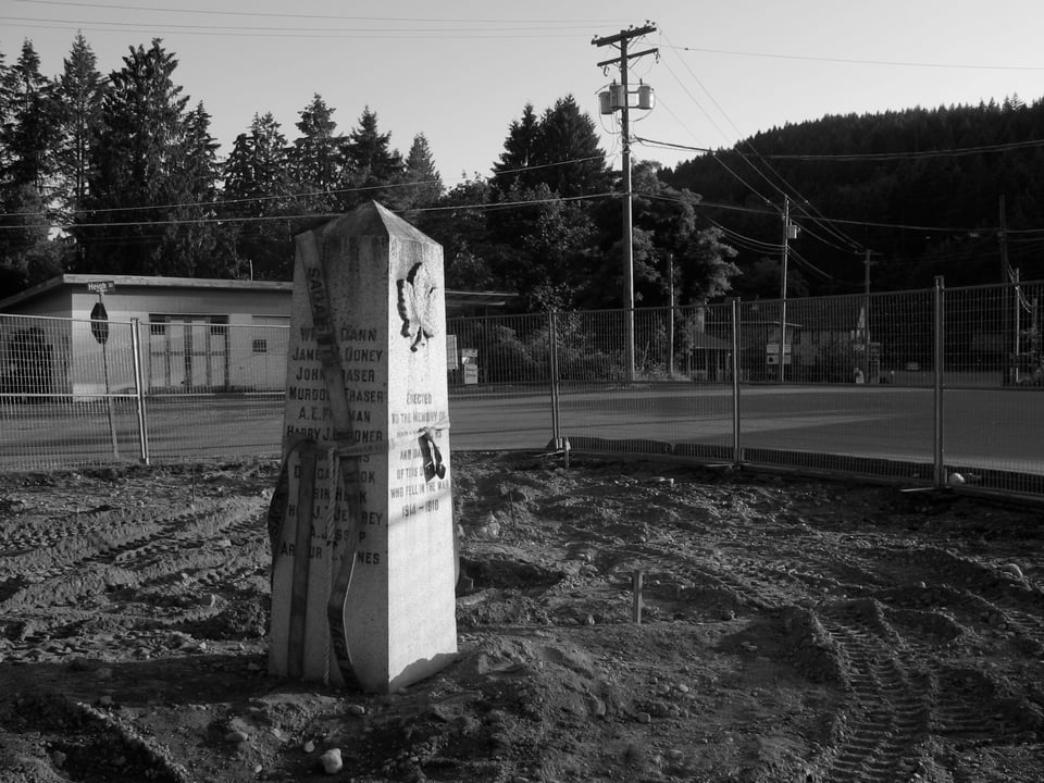 Black and white photo. A large granite obelisk marker with lettering stands in the middle of a dirt patch. There is fencing around the dirt patch and large straps for lifting are wrapped around the obelisk