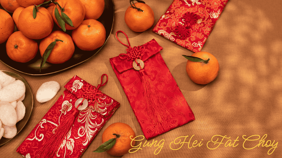 On a dappled beige surface: a dark ceramic plate of mandarin oranges, some with stems and leaves; a smaller plate of white sweets; three ornate red envelopes with patterned silk outer coverings and round jade clasps for red silk string enclosures with embroidered tassels. "Gung Hei Fat Choy" in script at lower right.