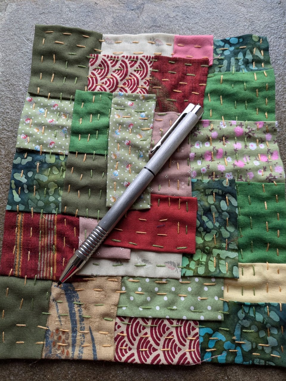 A colourful patchwork quilt about the size of a napkin, with a pen for scale