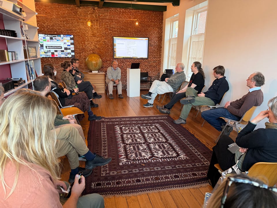 A group of people sit around a living room listening to a man talk as he sits next to a podium with a screen behind him.