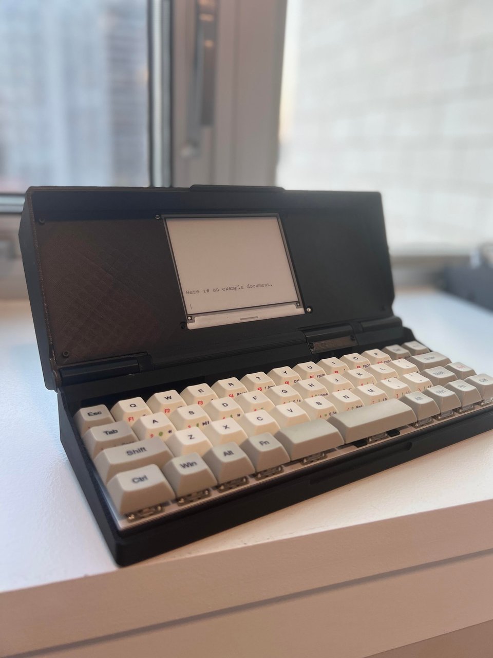 The Zerowriter, sitting on a white table. It is open. The keyboard is made of large white and grey thick chunky keys; the lid has a small square eink display perhaps 3 inches square, located in the center of the lid. The lid is black