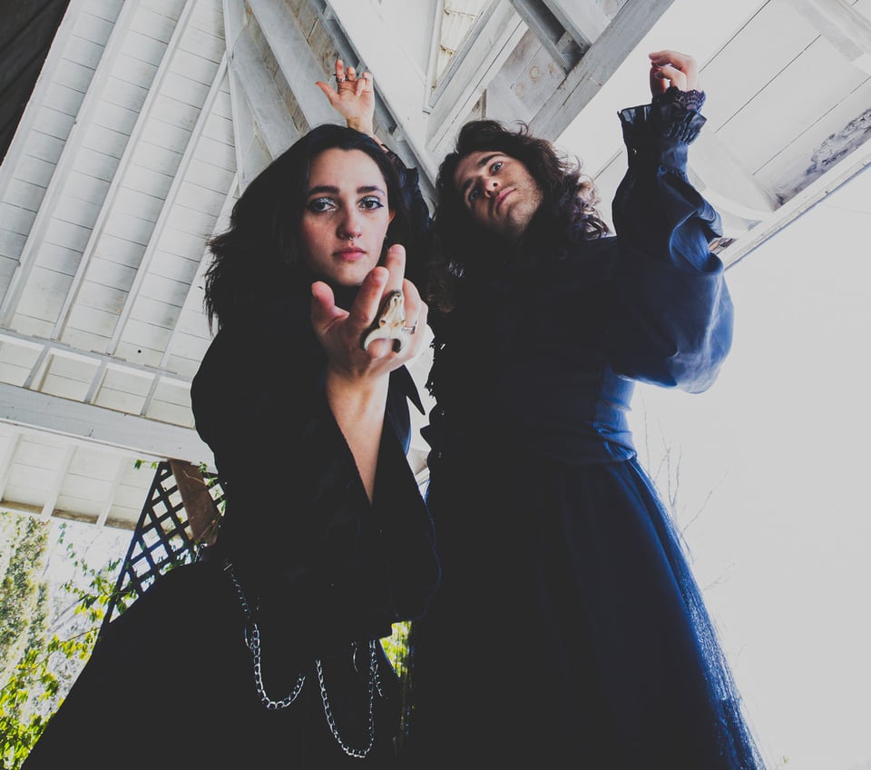 A woman with long dark hair leans toward the camera, her right hand extended as if beckoning the viewer. She stands next to a man with long hair and his arms upraised.