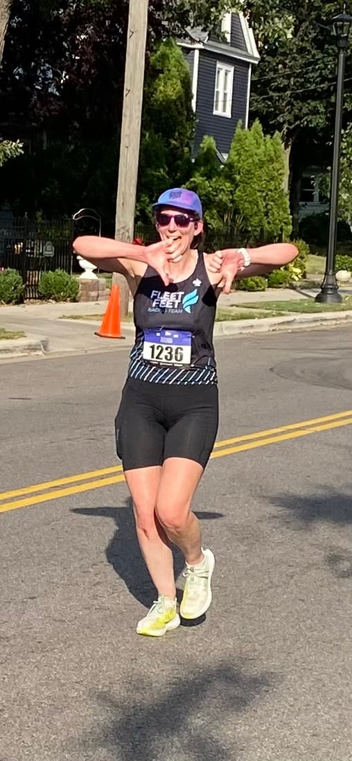 A woman running in a race (wearing black shorts, black and blue tank, white shoes, purple hat, purple glasses) showing both thumbs turned down