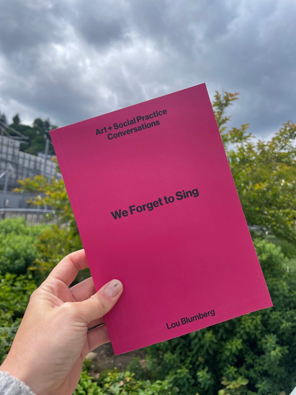 a hand holds up a pink book titled "We Forget To Sing" by Lou Blumberg on a green and grey background (plants and clouds).