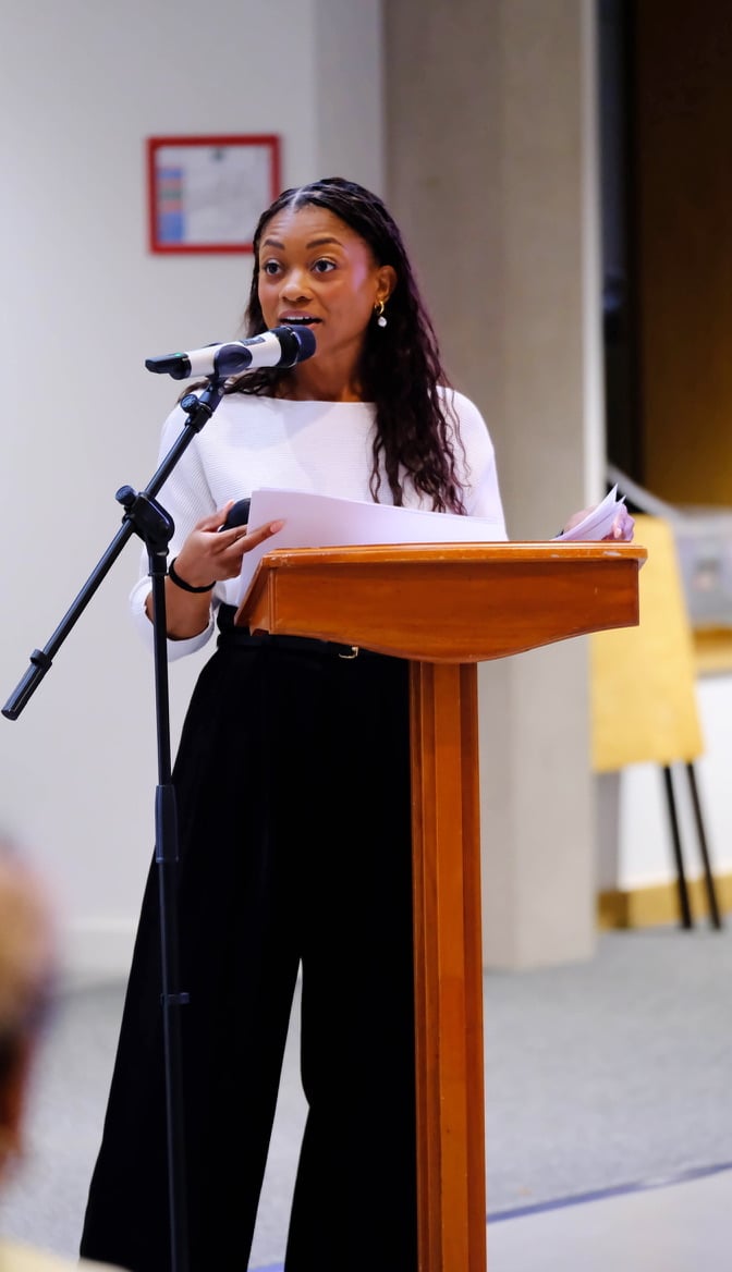 Nadia stands behind a wooden lectern, speaking into a microphone while holding sheets of paper. She wears a white long-sleeved top, black wide-leg trousers and gold earrings, with her long, wavy hair falling over her shoulders.