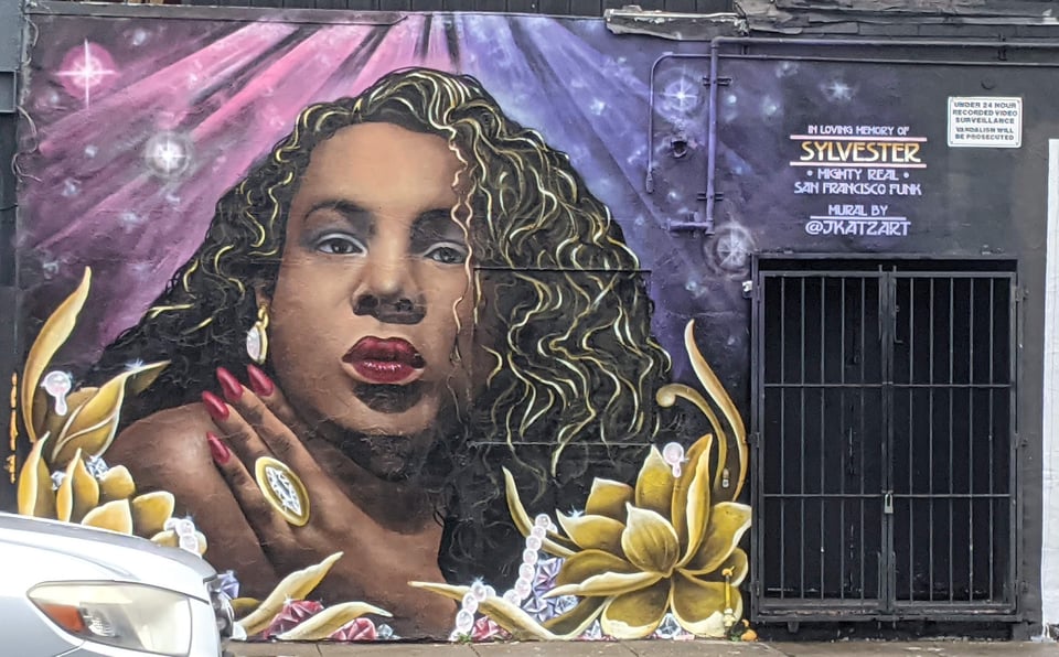 Sylvester, a feminine Black man with gorgeous makeup and long wavy hair with blonde streaks in it. He has long red nails and a gold ring, and he's surrounded by golden flowers and pearls.