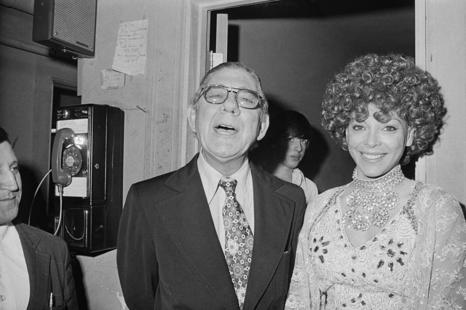 Wilbur Mills, an elderly congressman from Arkansas, wearing a loose tie with a lively pattern, next to Fanne Fox, who has a big curly wig and an elaborate necklace with an embroidered dress and shawl
