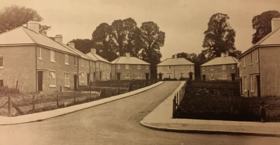 a sepia photo of a cul-de-sac of semidetached houses. They are blocky and square, with no decoration. The gardens are fenced with low chicken wire, and there is nothing in the road. There are large trees beyond the houses.